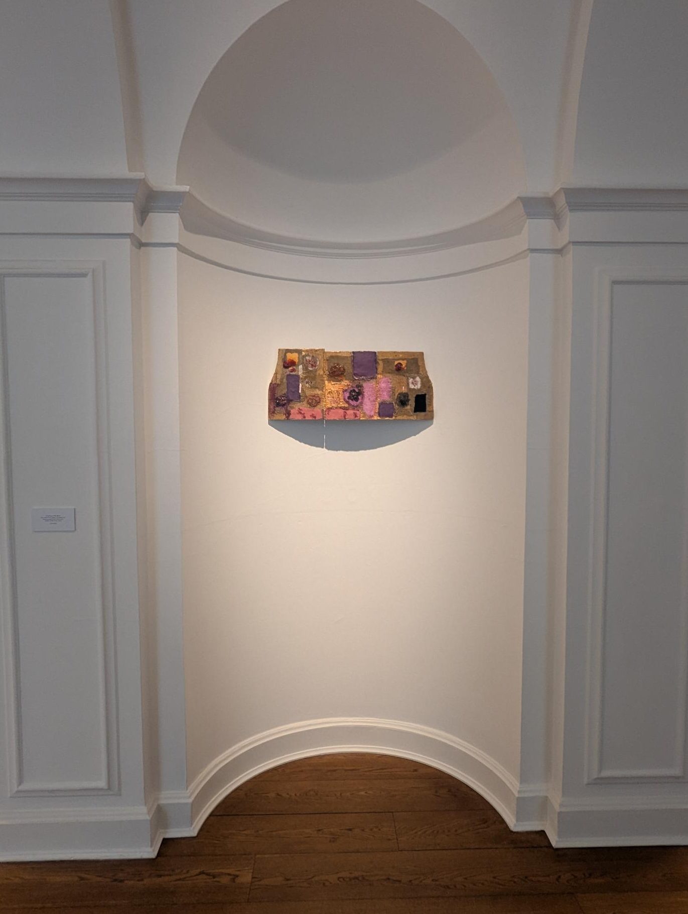An artwork by Joan Snyder displayed on a wall in an elegant gallery setting, featuring textured and colorful elements arranged in a wavy rectangular shape.